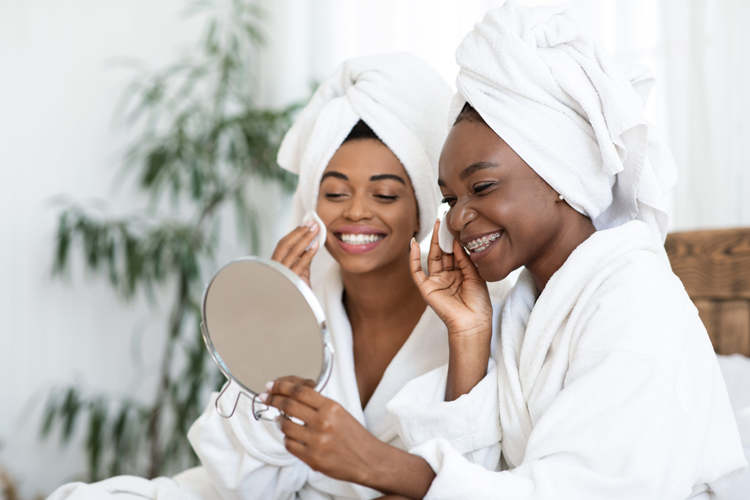 Attractive african american ladies applying toner on faces with cotton pads, looking at mirror. Prertty black women having beauty day at home, looking at mirror and cleansing their faces, copy space